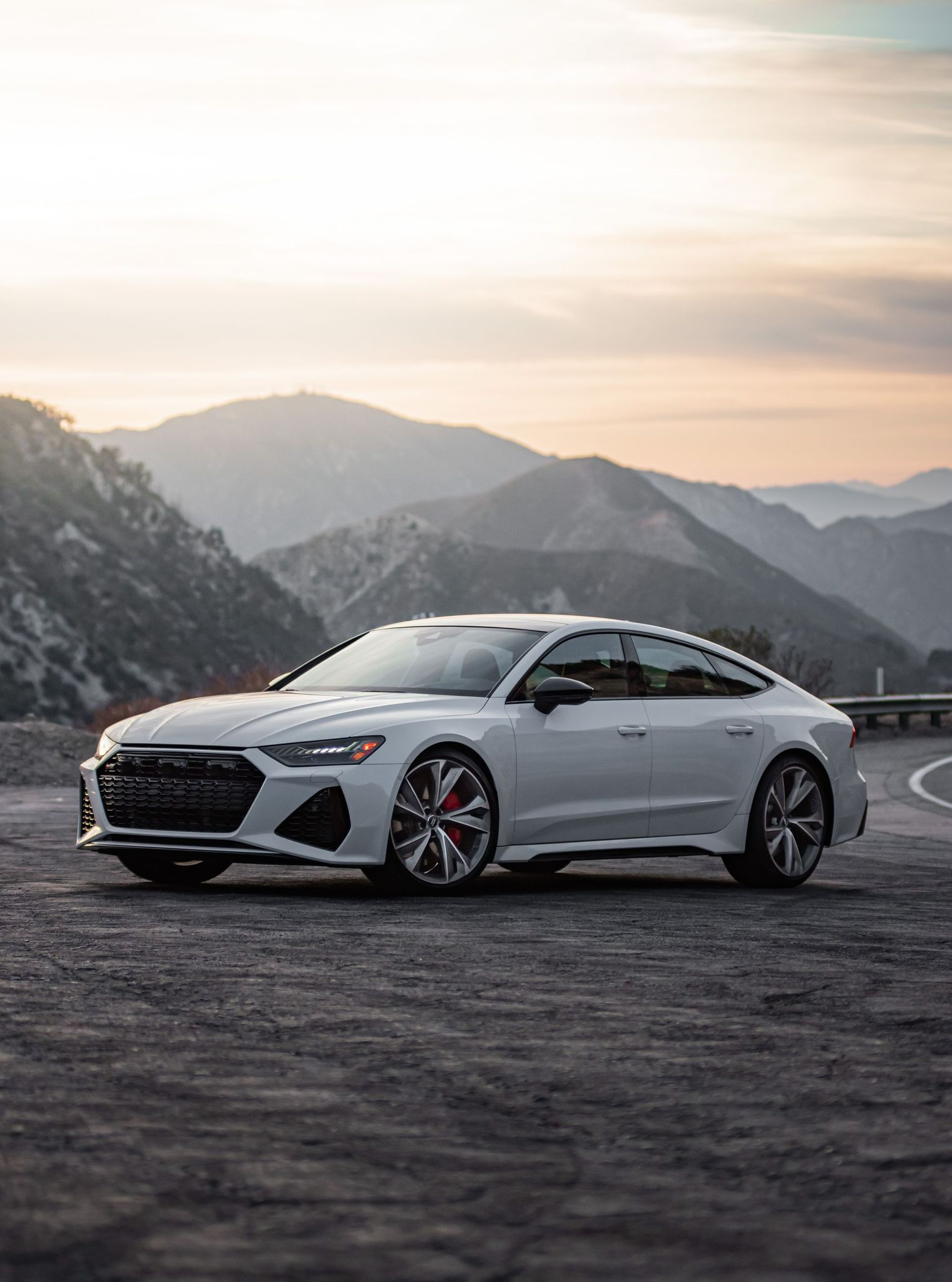 White Audi RS7 parked on a mountain road with a mountainous backdrop at sunset.