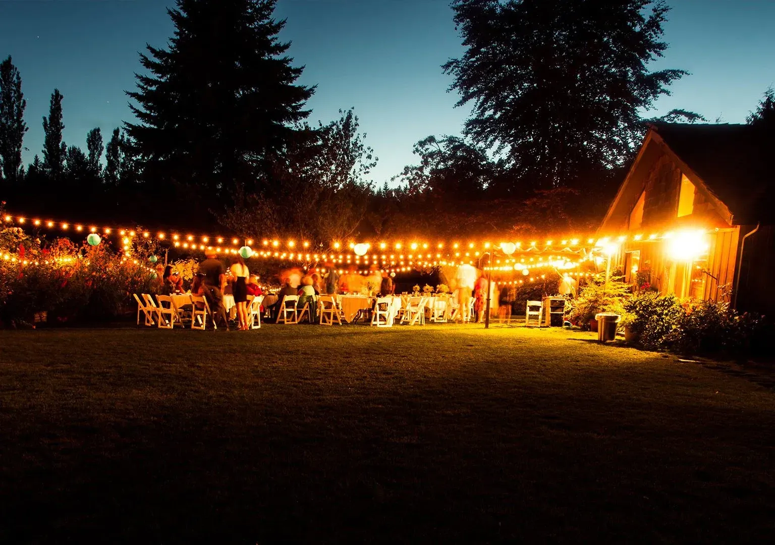 Een gezellige avondbijeenkomst in de buitenlucht bij lichtslingers in de buurt van een verlichte hut, met mensen die aan tafels op een gazon zitten.