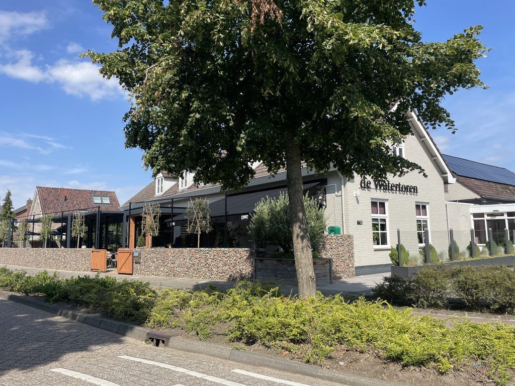 Modern gebouw met stenen muur, grote boom en aangelegde berm onder een blauwe hemel.