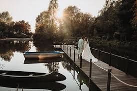 Een stel in witte kleding op een steiger boven kalm water bij zonsondergang, met een boot op de voorgrond.
