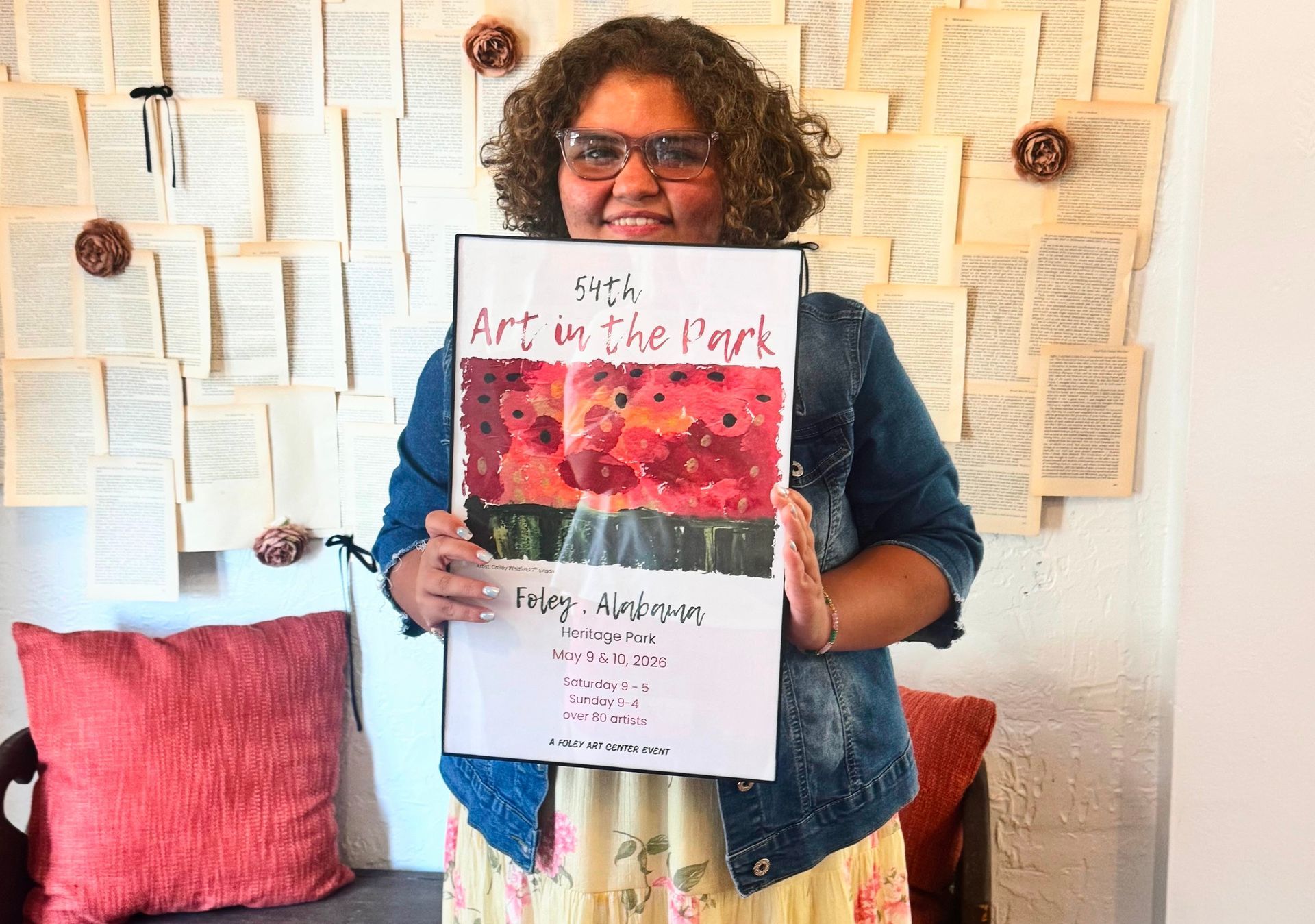 Cailey Whitfield holds the 54th Annual Art in the Park poster featuring her artwork, depicting a bouquet of flowers in vivid shades of red, purple, and pink.