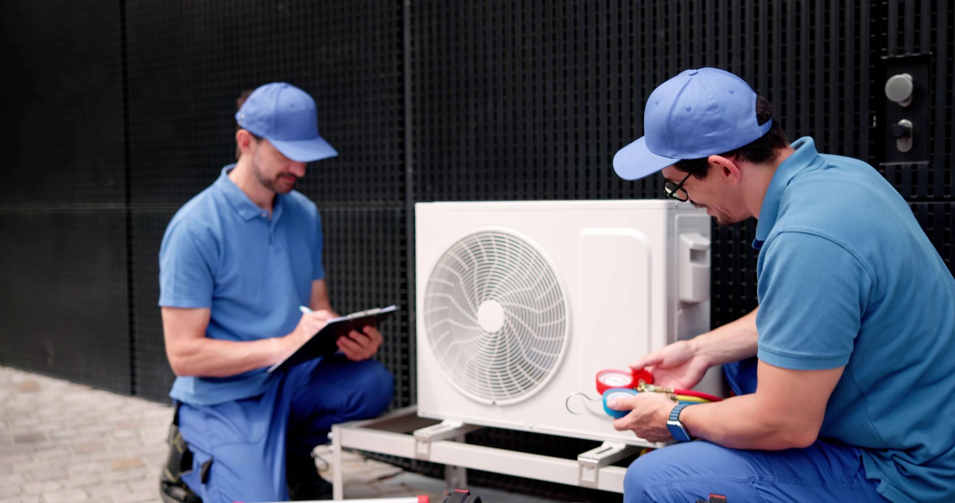 Two HVAC technicians in blue uniforms work on an air conditioning unit. One checks a clipboard, the other attaches gauges. Two HVAC technicians in blue uniforms work on an air conditioning unit. One checks a clipboard, the other attaches gauges.