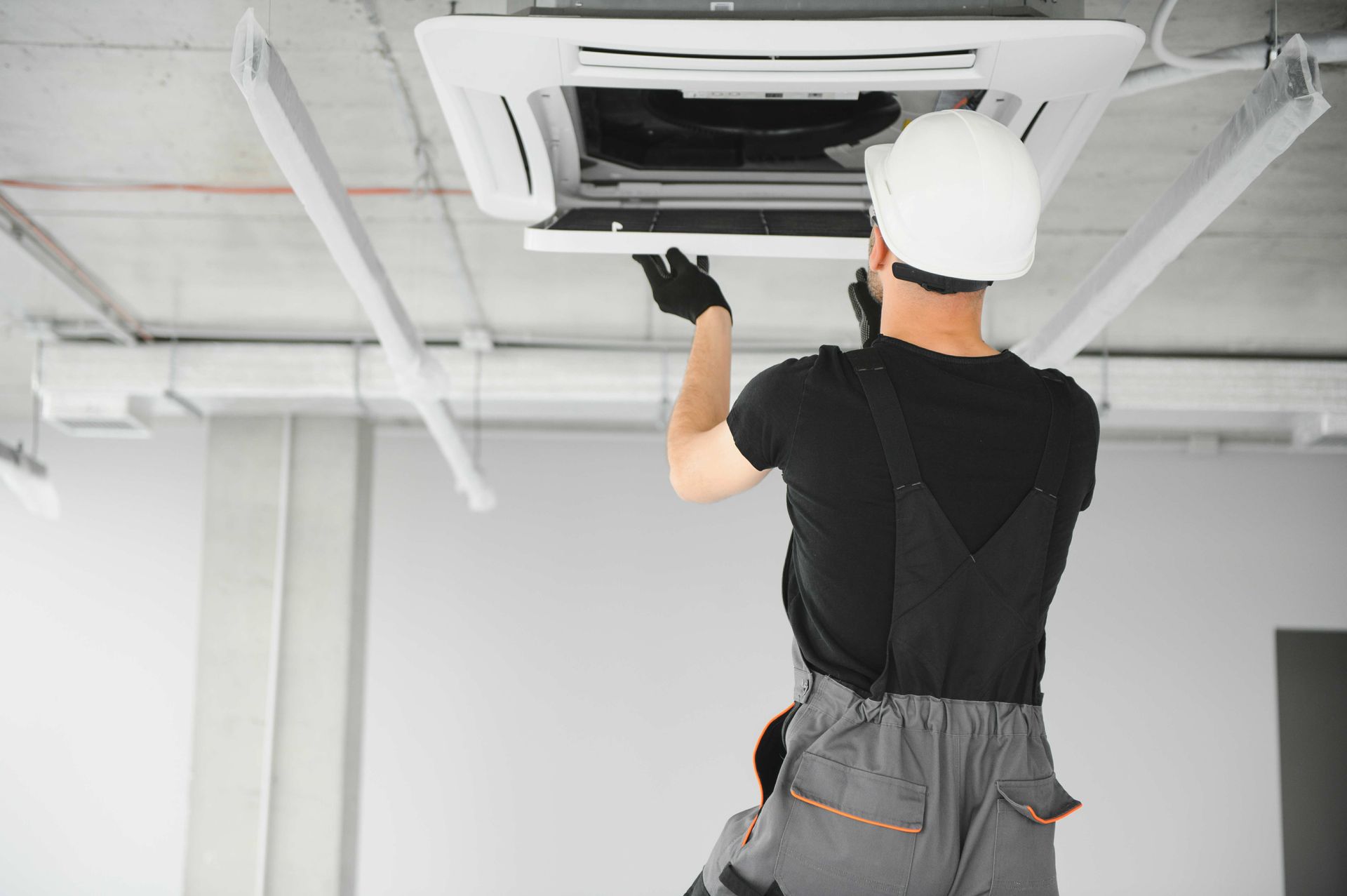 HVAC technician in overalls, hard hat, and gloves, repairing ceiling air conditioner in a building.