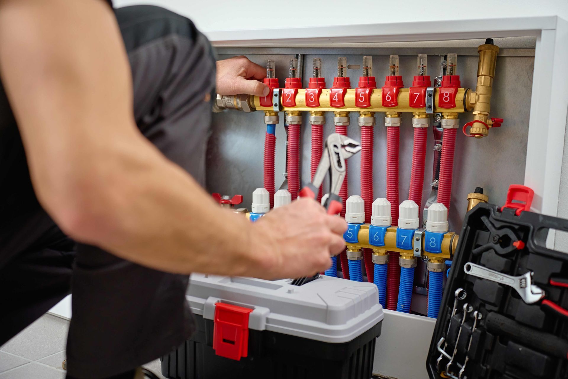 Plumber using pliers to work on a radiant floor heating manifold in a white utility space. Plumber using pliers to work on a radiant floor heating manifold in a white utility space.