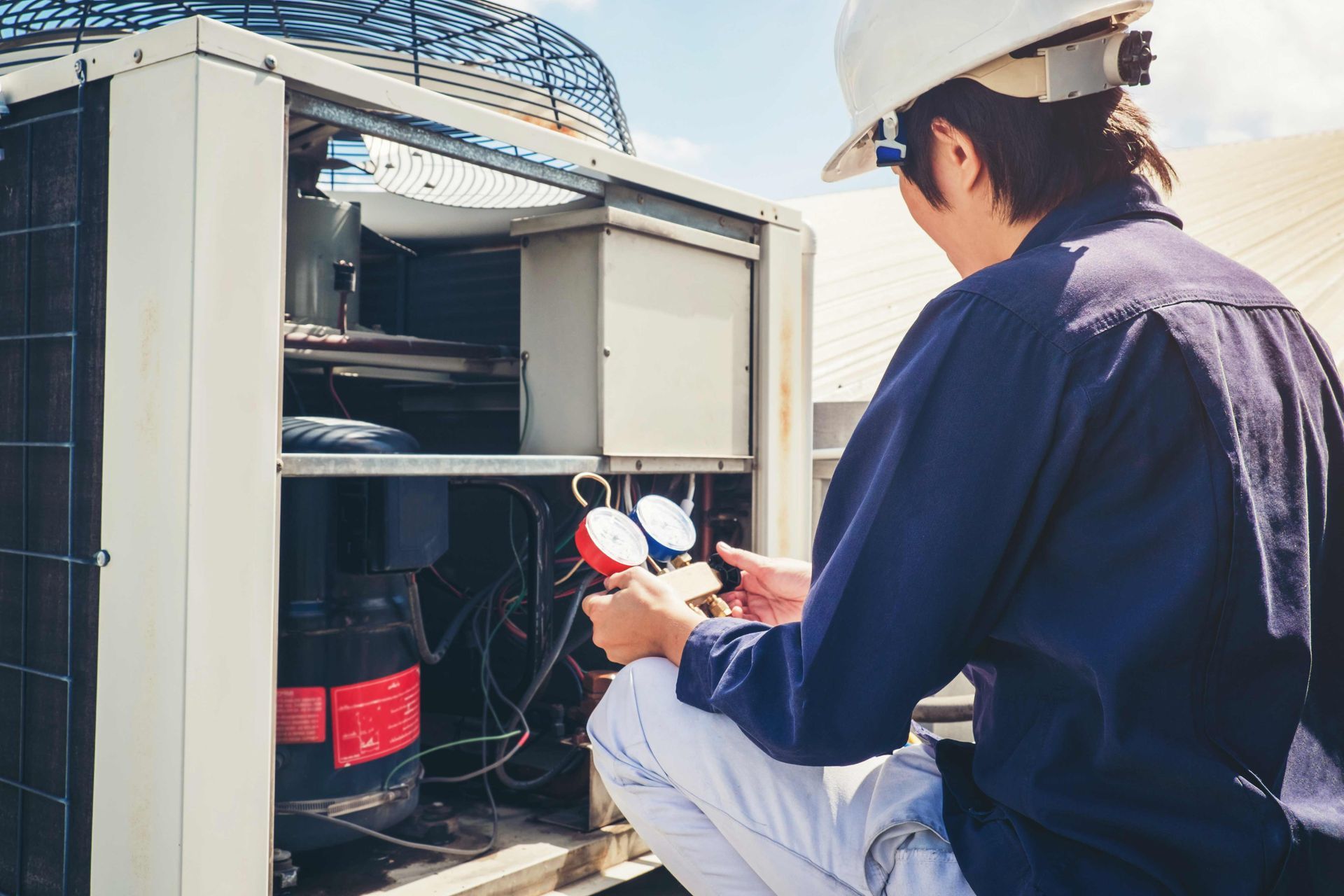 HVAC technician kneeling near open AC unit on rooftop, holding gauges to diagnose.
