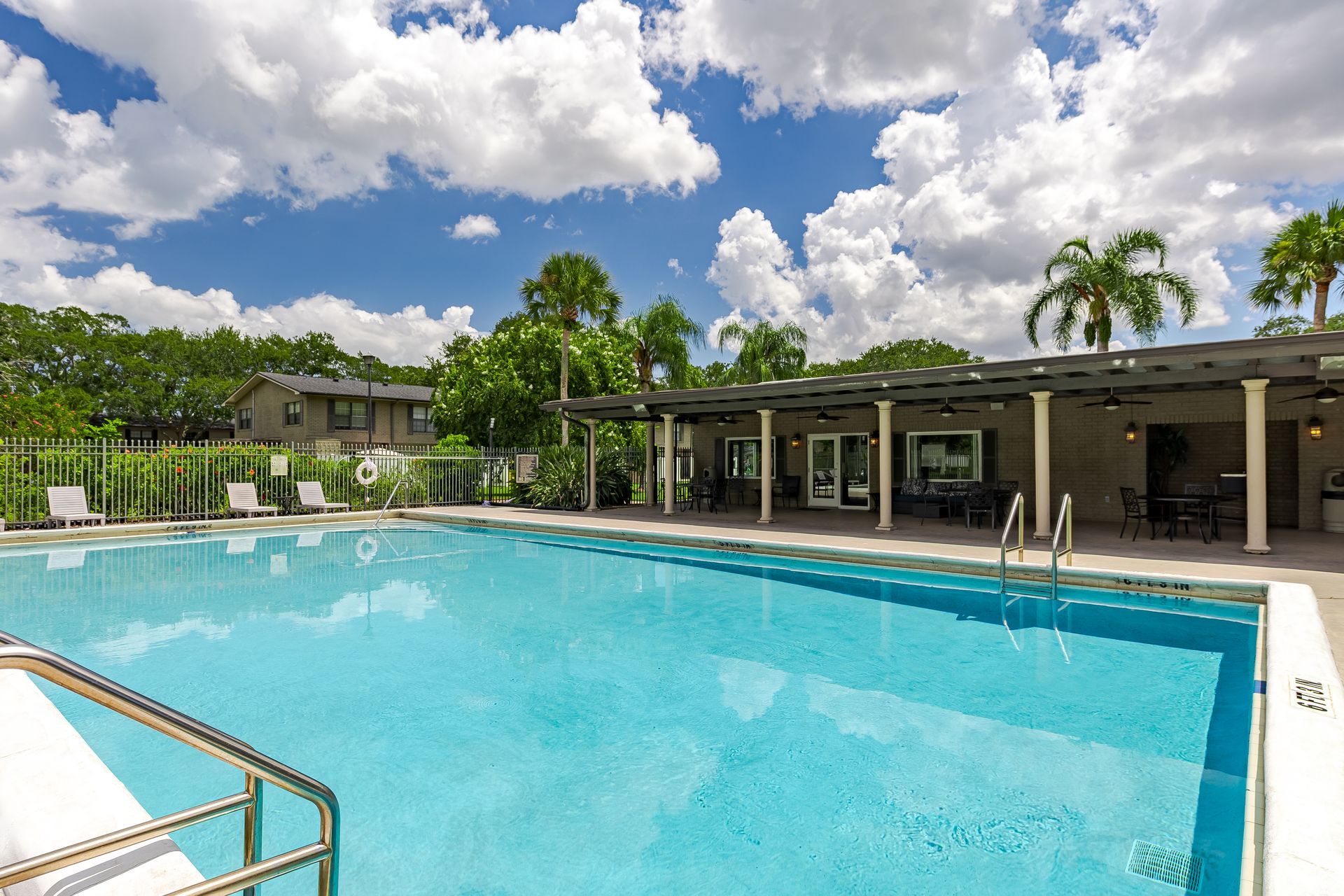 A clear, rectangular swimming pool with a covered patio area and palm trees against a blue sky with white clouds at Veridian of Melbourne Townhome Apartments in Melbourne, FL.