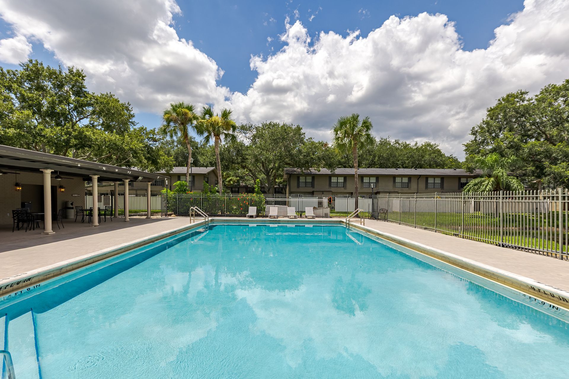 An outdoor swimming pool with bright blue water, surrounded by a patio, palm trees, and multi-story apartment buildings at Veridian of Melbourne Townhome Apartments in Melbourne, FL.