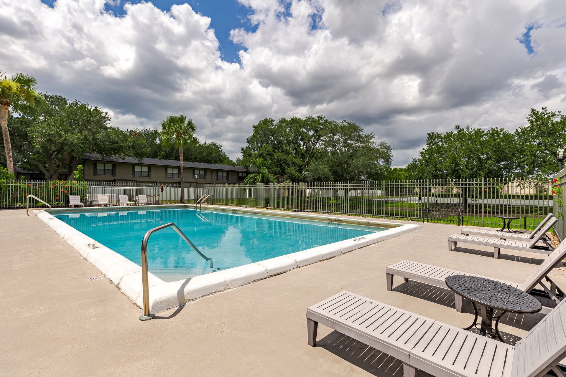 A rectangular swimming pool surrounded by lounge chairs on a concrete deck, with palm trees and a building in the distance at Veridian of Melbourne Townhome Apartments in Melbourne, FL.