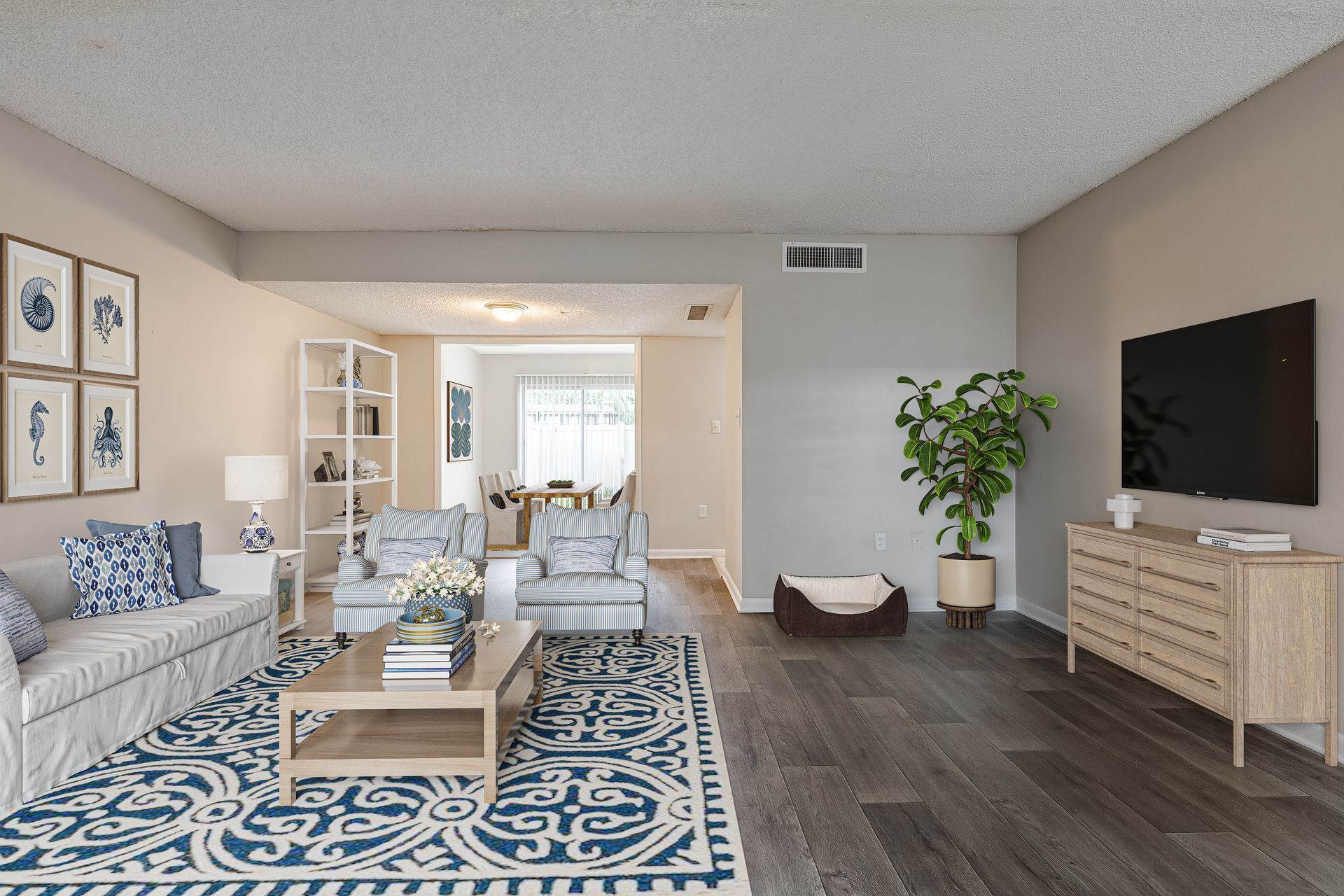 A living room with a white sofa, blue patterned rug, two armchairs, a wooden coffee table, TV, and a green potted plant at Veridian of Melbourne Townhome Apartments in Melbourne, FL.