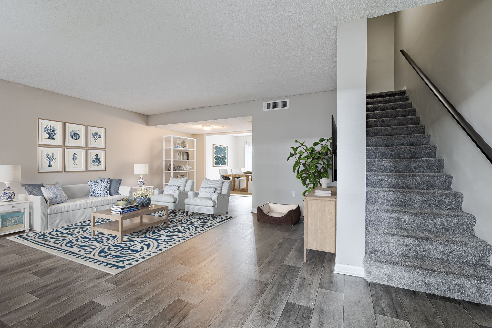 Living room with light gray walls, wooden floors, blue area rug, seating, and a gray carpeted staircase on the right at Veridian of Melbourne Townhome Apartments in Melbourne, FL.