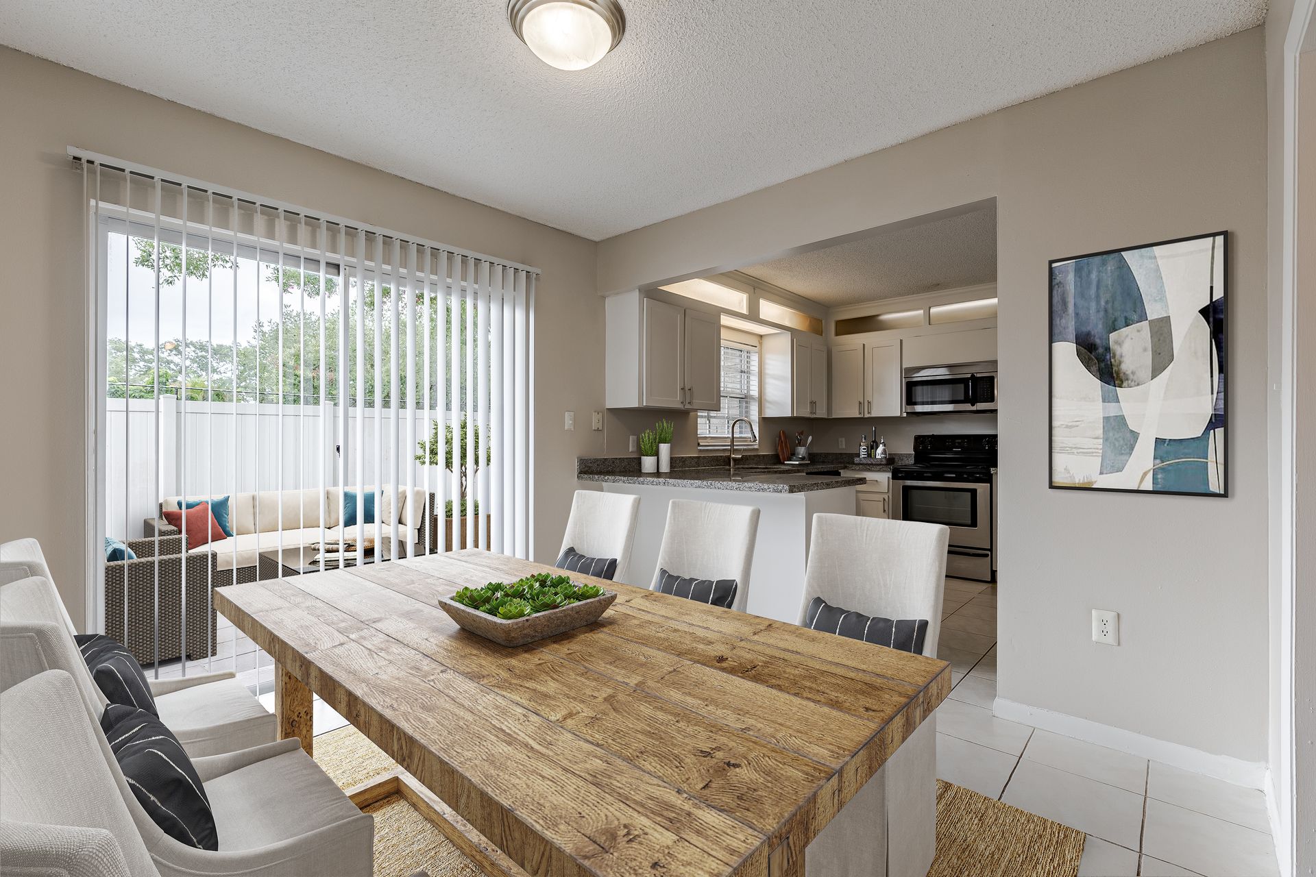 A dining room with a wooden table, white chairs, and a sliding glass door opening to a patio, with a kitchen in the back at Veridian of Melbourne Townhome Apartments in Melbourne, FL.
