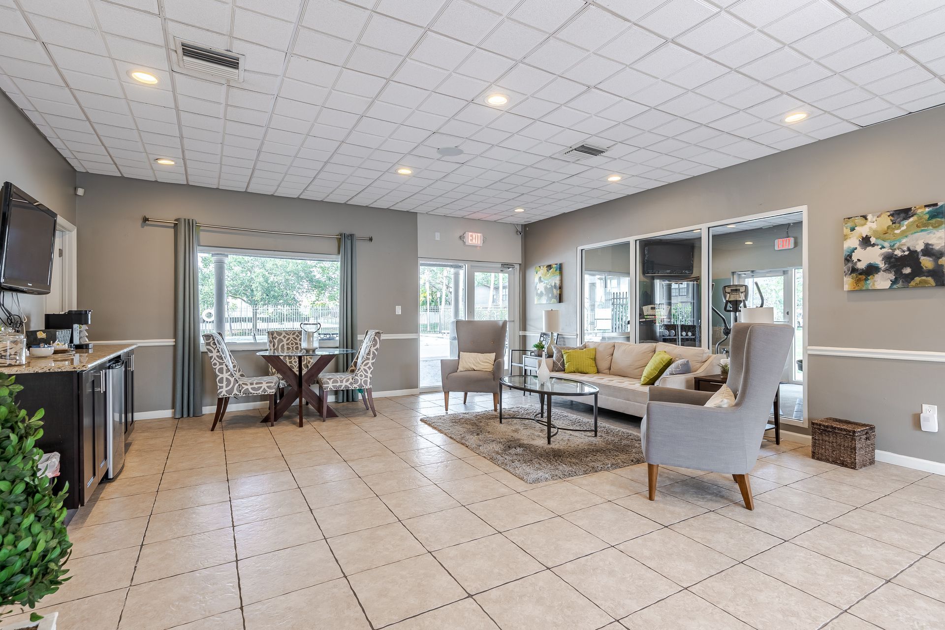A well-lit, contemporary apartment clubhouse lounge featuring a sofa, gray armchairs, a dining set, and tiled flooring at Veridian of Melbourne Townhome Apartments in Melbourne, FL.