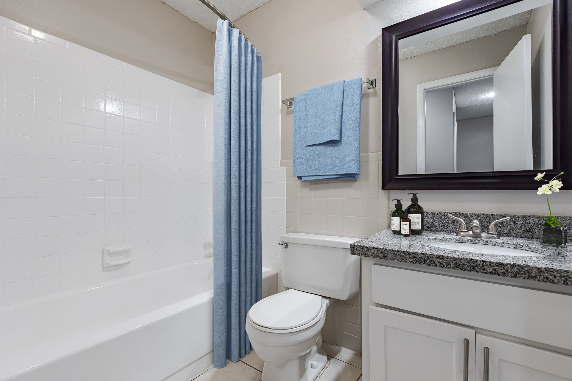 A modern bathroom featuring a white tub with a blue curtain, a white toilet, and a vanity with a granite top and mirror at Veridian of Melbourne Townhome Apartments in Melbourne, FL.