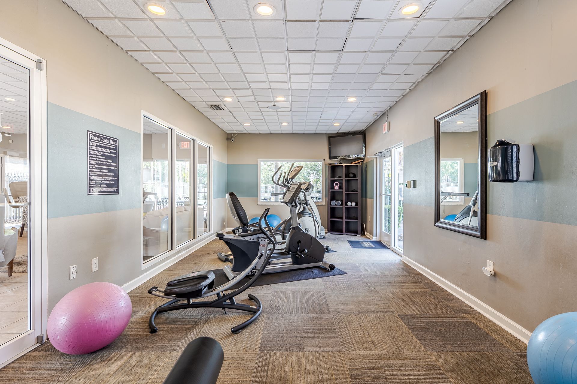 A bright, carpeted gym featuring exercise bikes, yoga balls, a television, and a wall mirror at Veridian of Melbourne Townhome Apartments in Melbourne, FL.