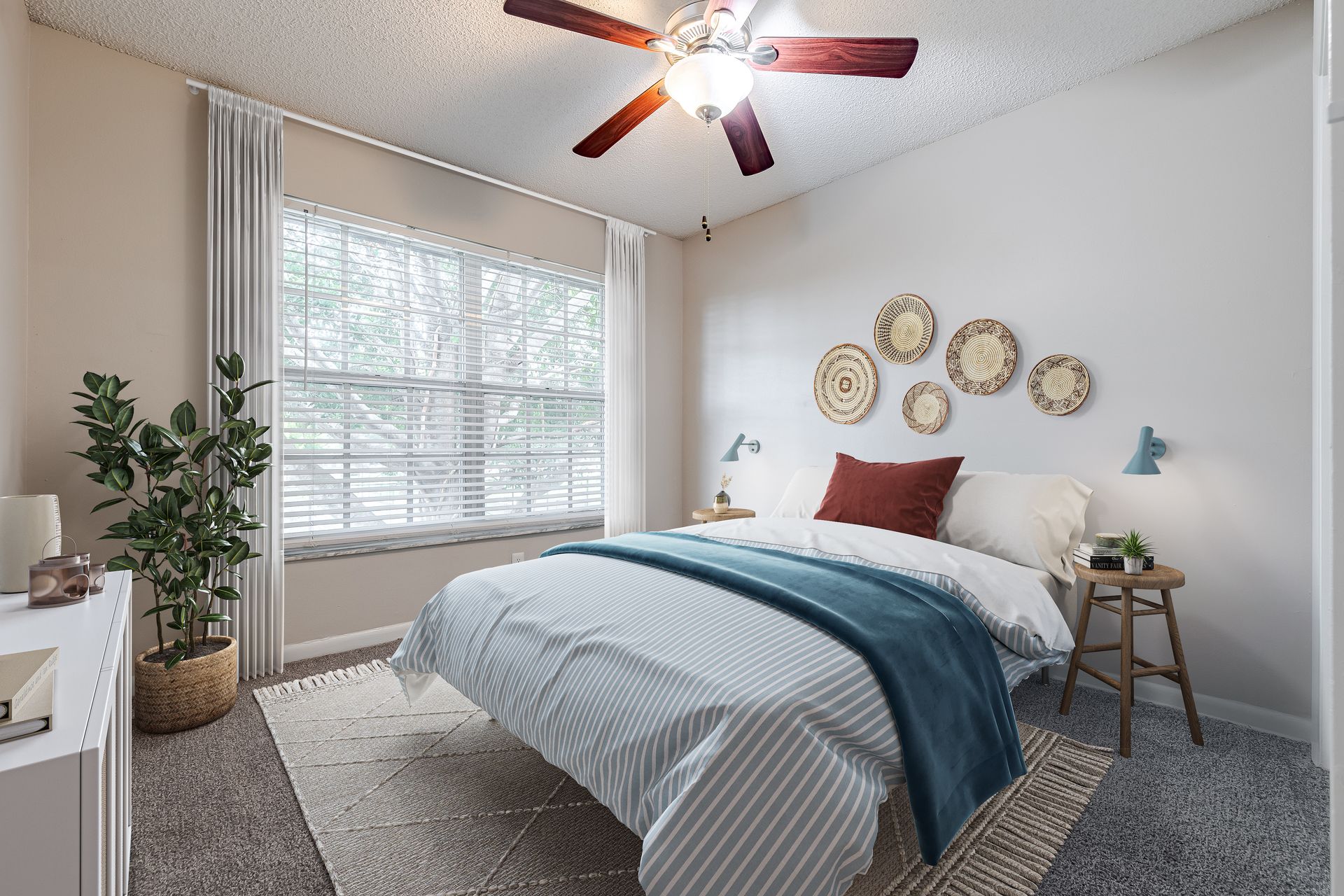 A well-lit bedroom featuring a queen bed with blue and white linens, a large window, a ceiling fan, and wall decor at Veridian of Melbourne Townhome Apartments in Melbourne, FL.