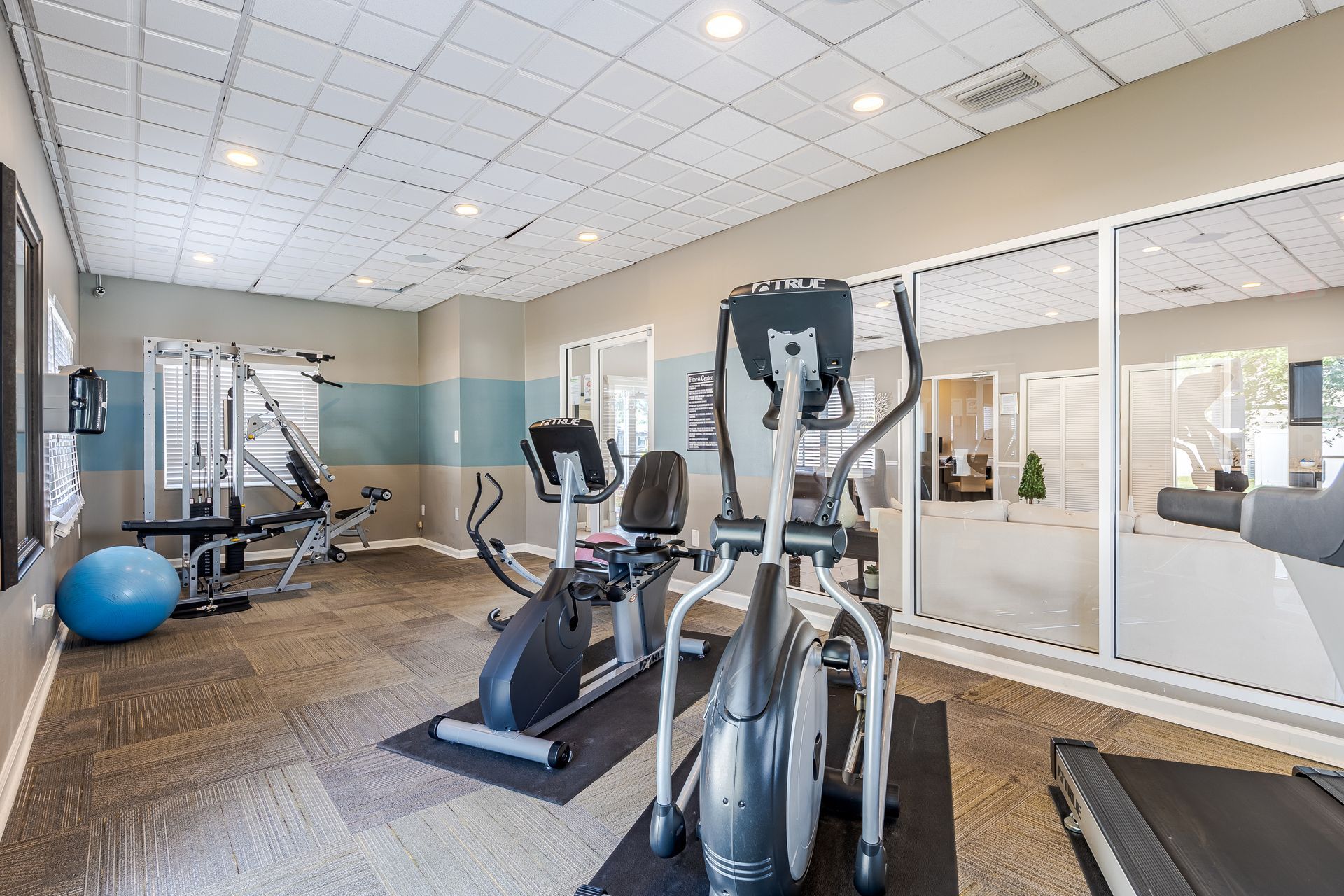 A bright, well-lit apartment fitness center featuring a stationary bike, elliptical, weight machine, and exercise ball at Veridian of Melbourne Townhome Apartments in Melbourne, FL.