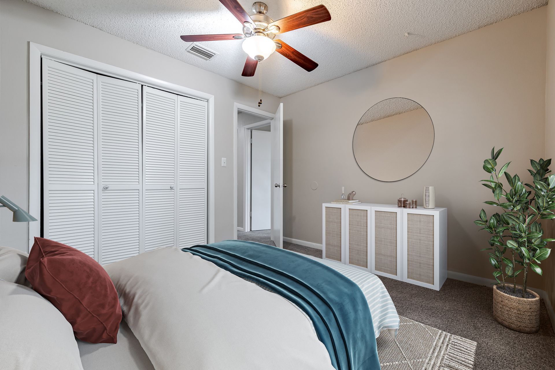 A bedroom featuring a bed with a blue throw, white closet doors, a round wall mirror, and a potted plant at Veridian of Melbourne Townhome Apartments in Melbourne, FL.