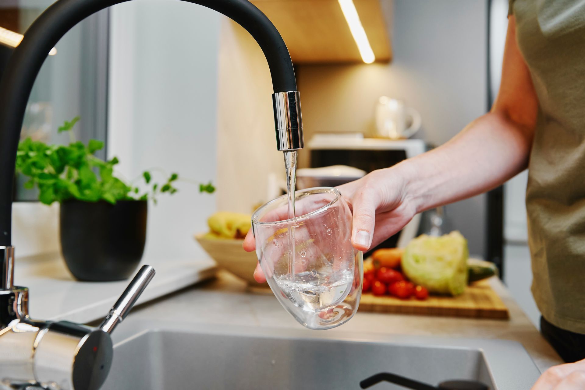 Person filling a glass with water from a kitchen faucet near a sink and cutting board