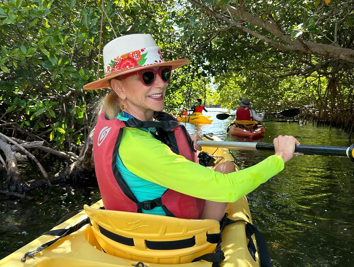 Woman kayaking in a mangrove tunnel, wearing a hat, sunglasses, and life vest.