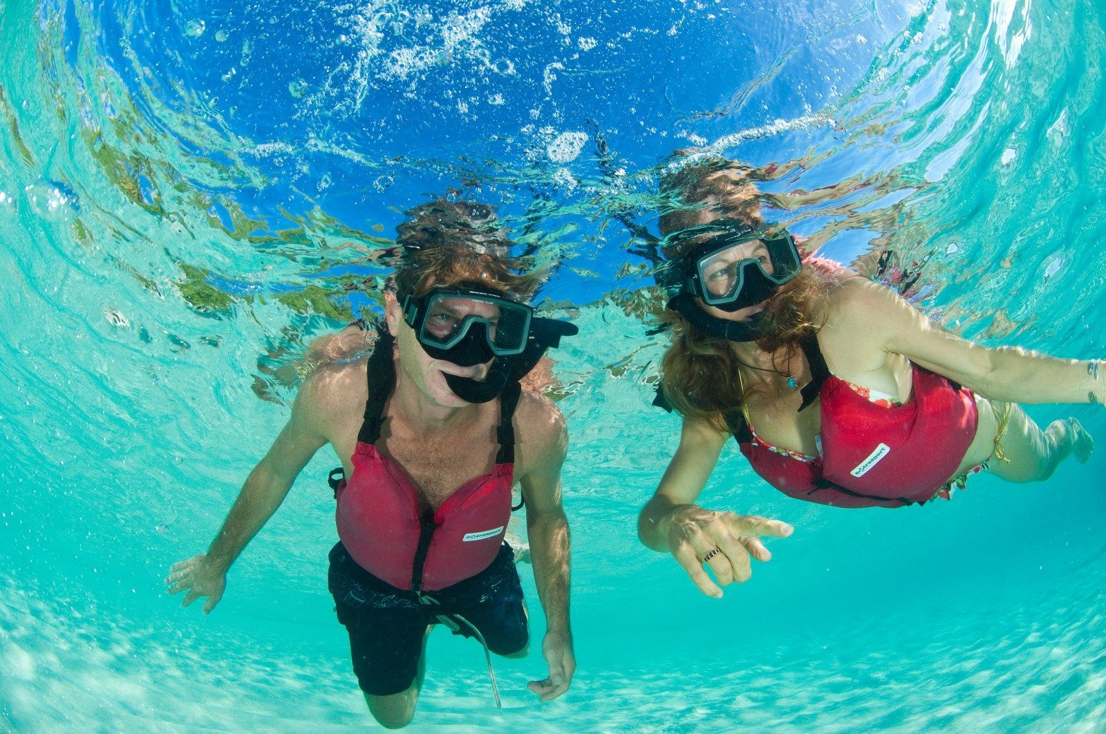 Two people snorkeling in clear blue water. They wear masks and red life vests.