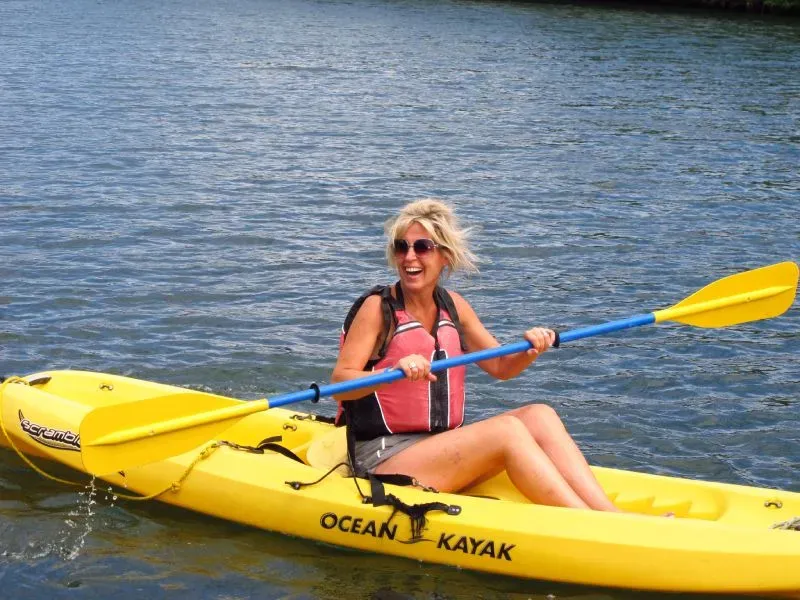 Woman kayaking in yellow kayak; blue paddle, life vest, smiling, on water.