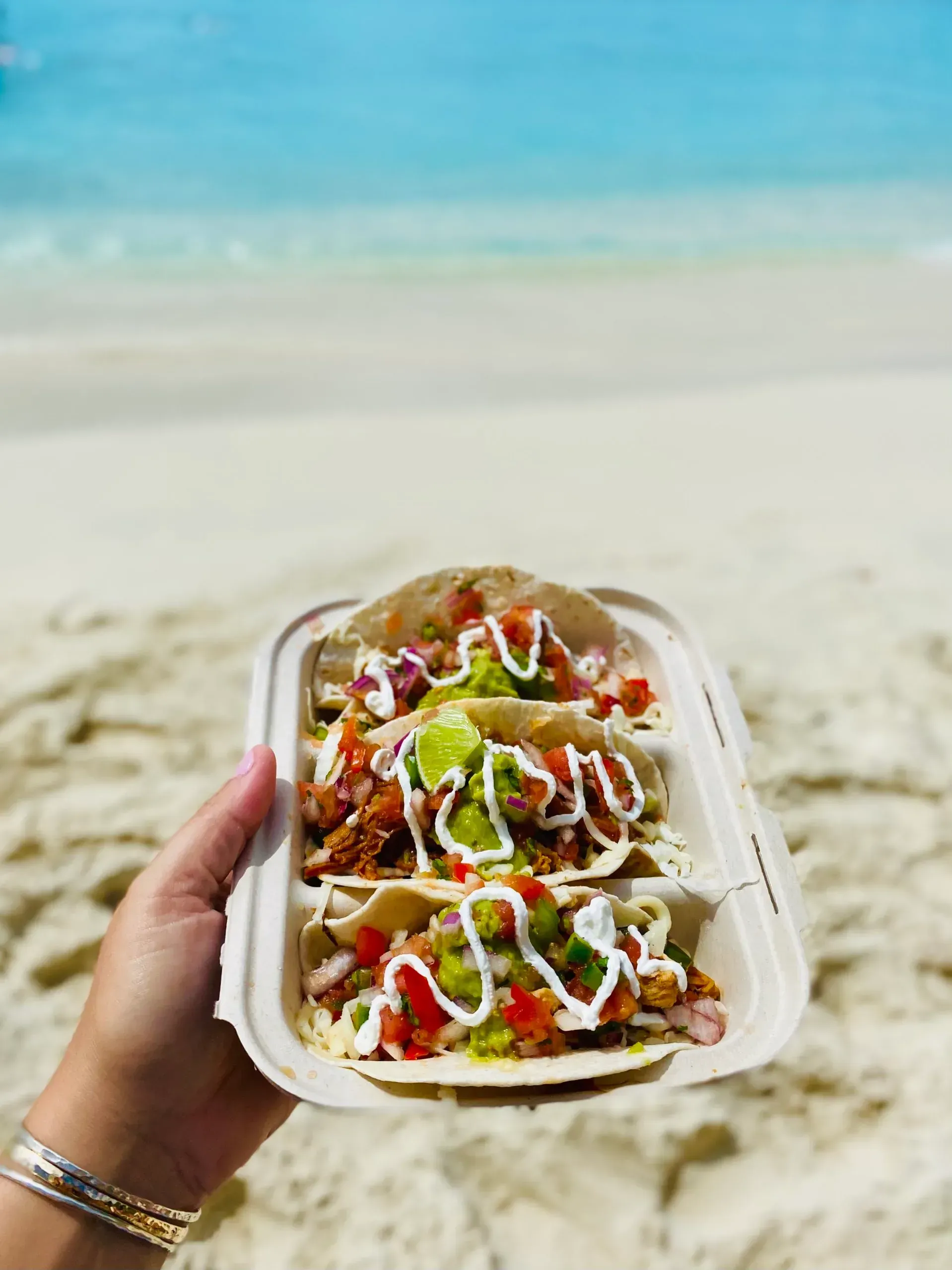 A person is holding a tray of tacos on the beach.
