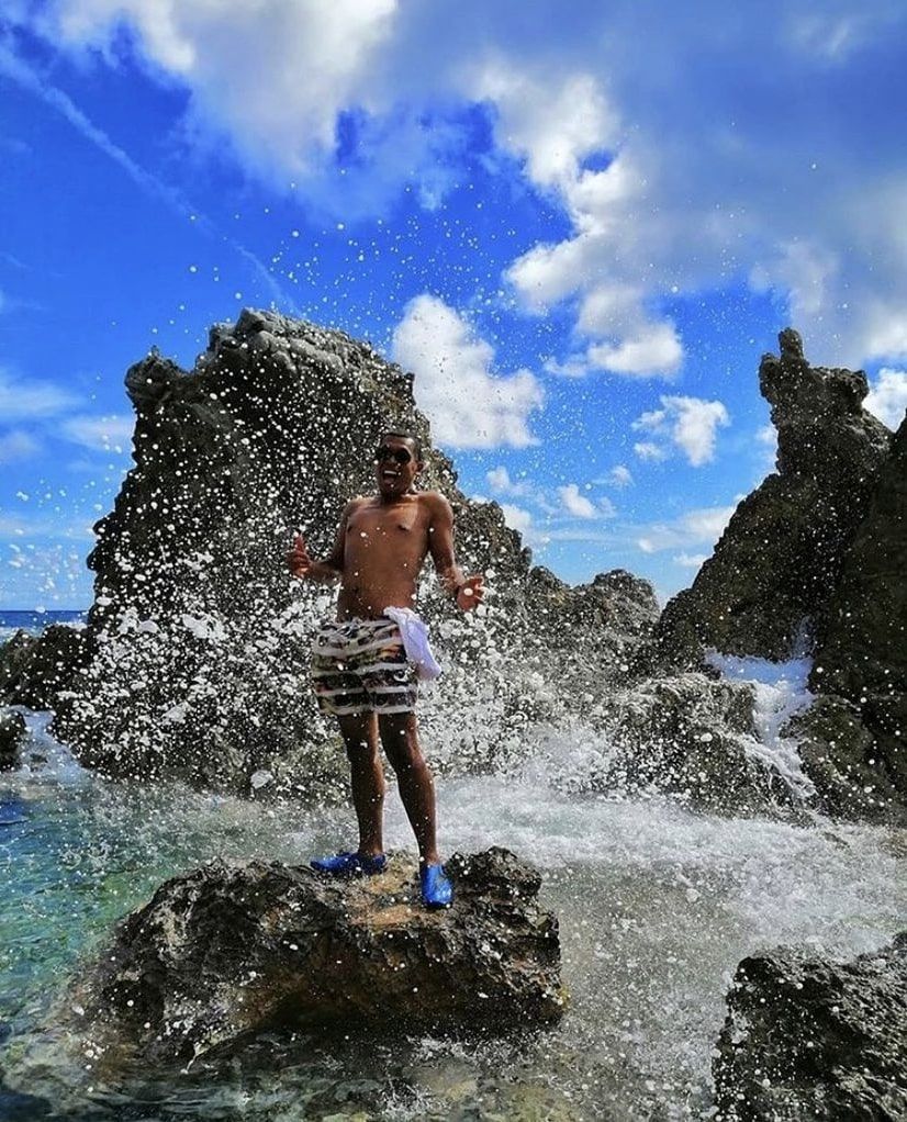 A shirtless man is standing on a rock in the ocean splashing water.