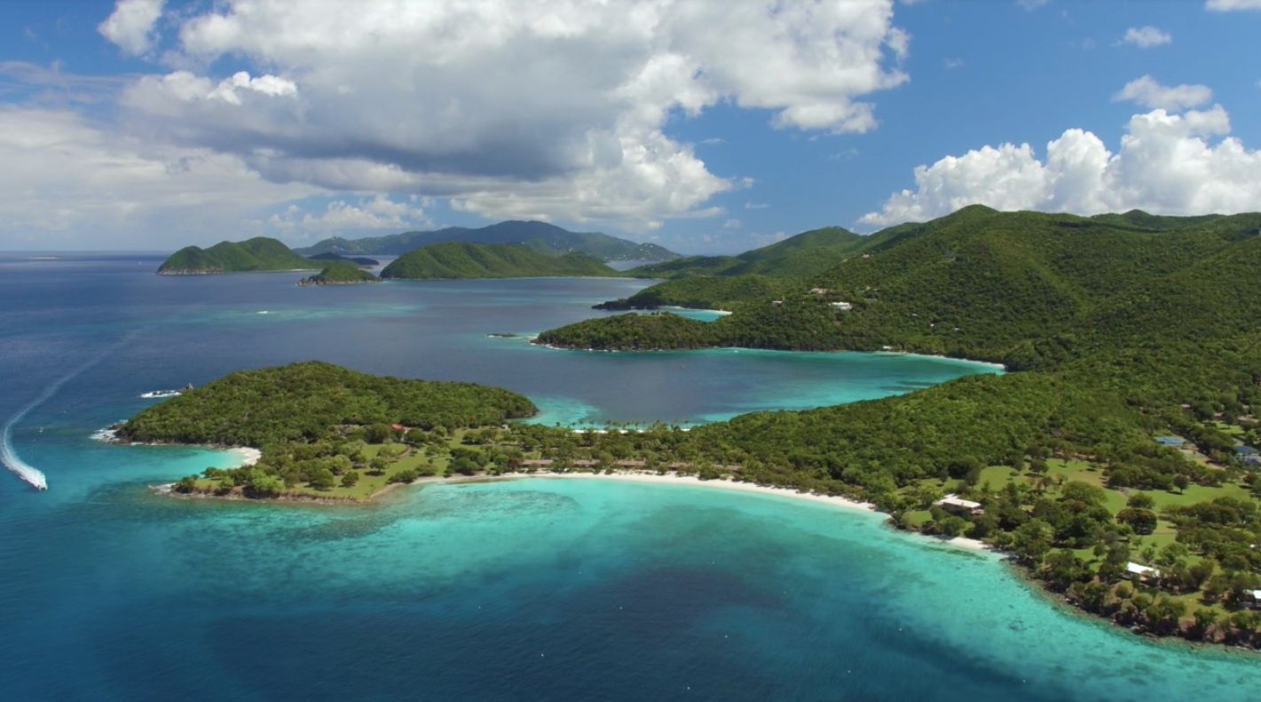 An aerial view of a tropical island with turquoise water and lush green vegetation under a partly cloudy sky.