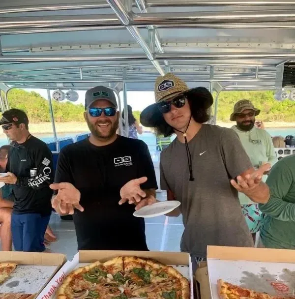 A man wearing sunglasses and a hat is standing next to a box of pizza