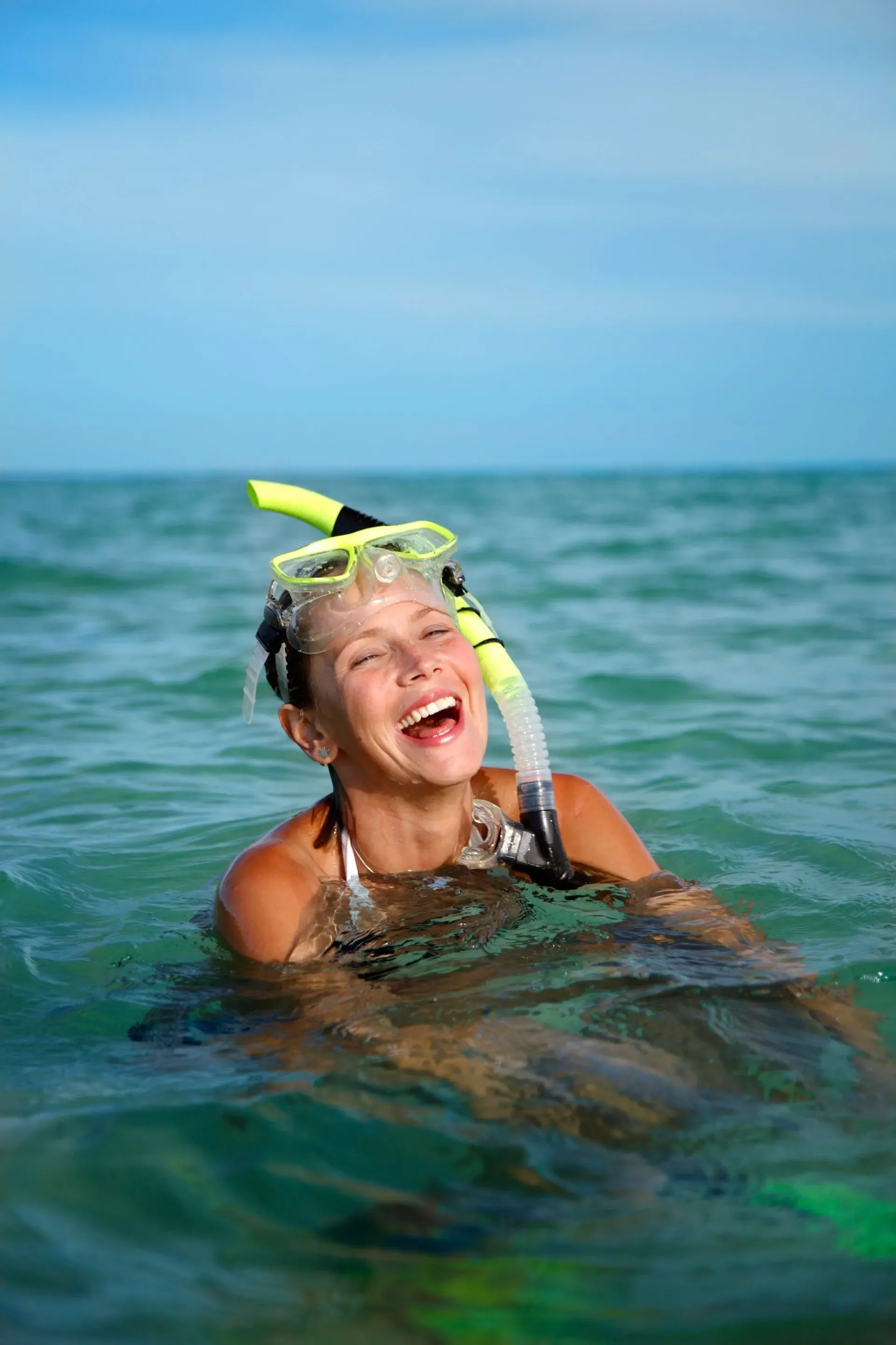 Woman in water, laughing, wearing snorkel gear. Blue sky and sea in background.