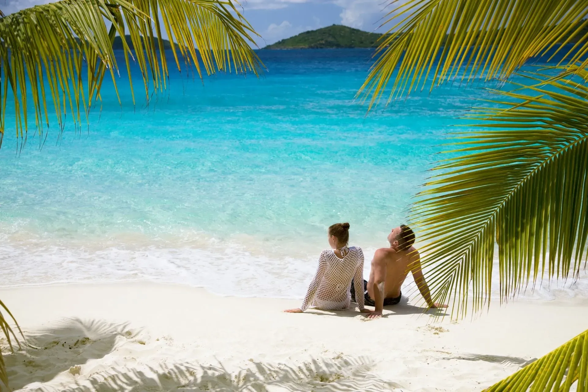 Couple relaxing on white sand beach, turquoise water, palm trees.