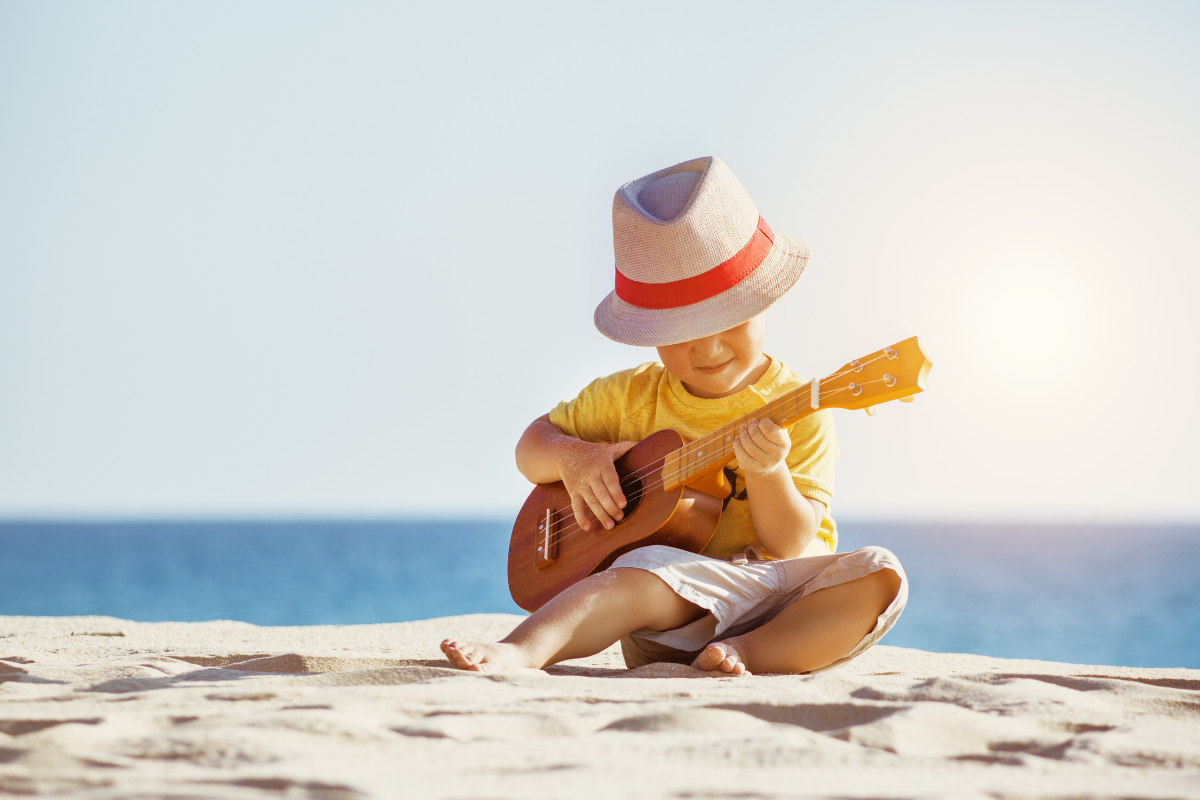A young boy is sitting on the beach playing a guitar.