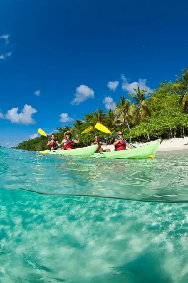 Kayakers paddling in turquoise water near a tropical beach under a blue sky.
