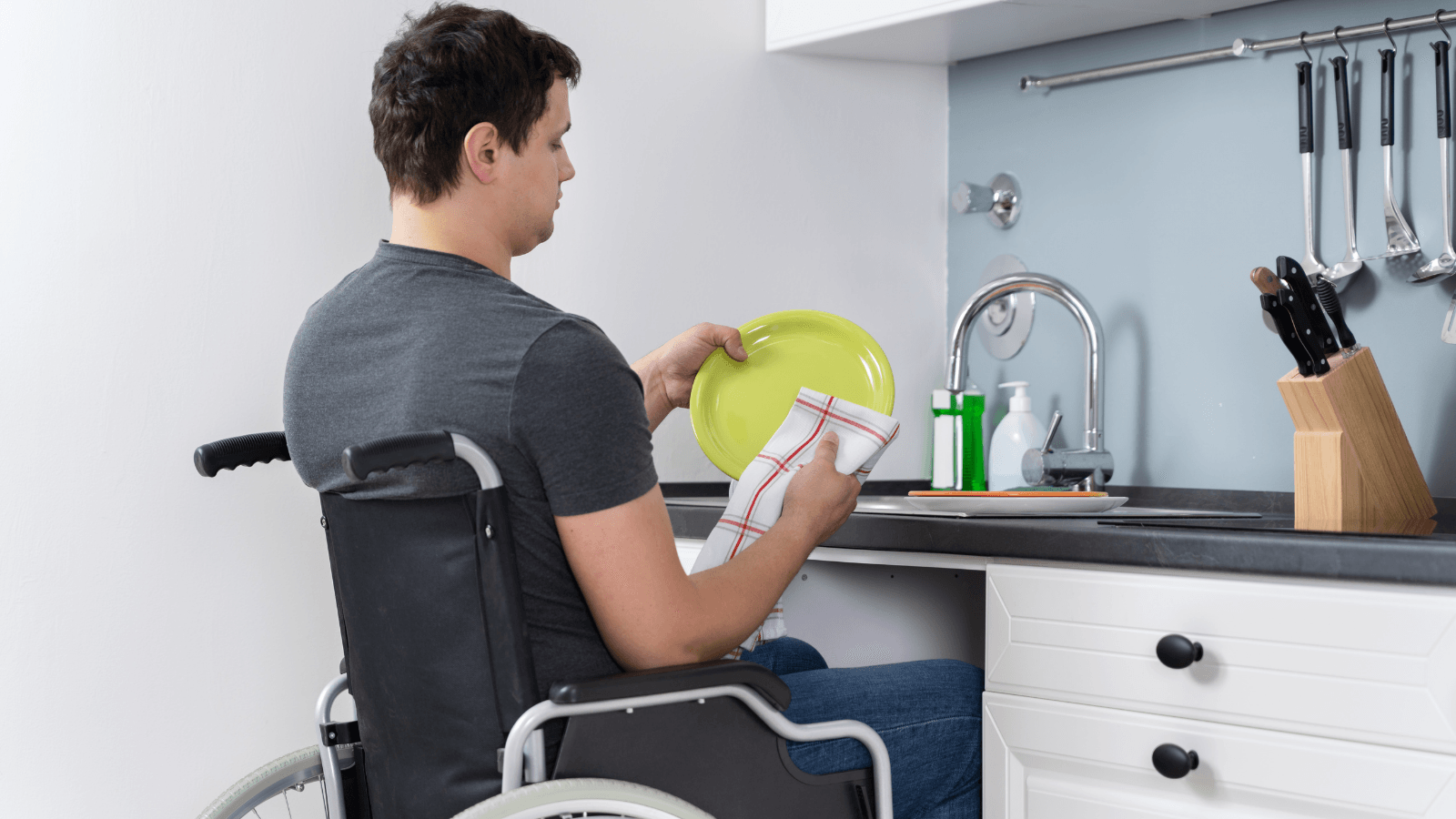 A man in a wheelchair is washing dishes in the kitchen.