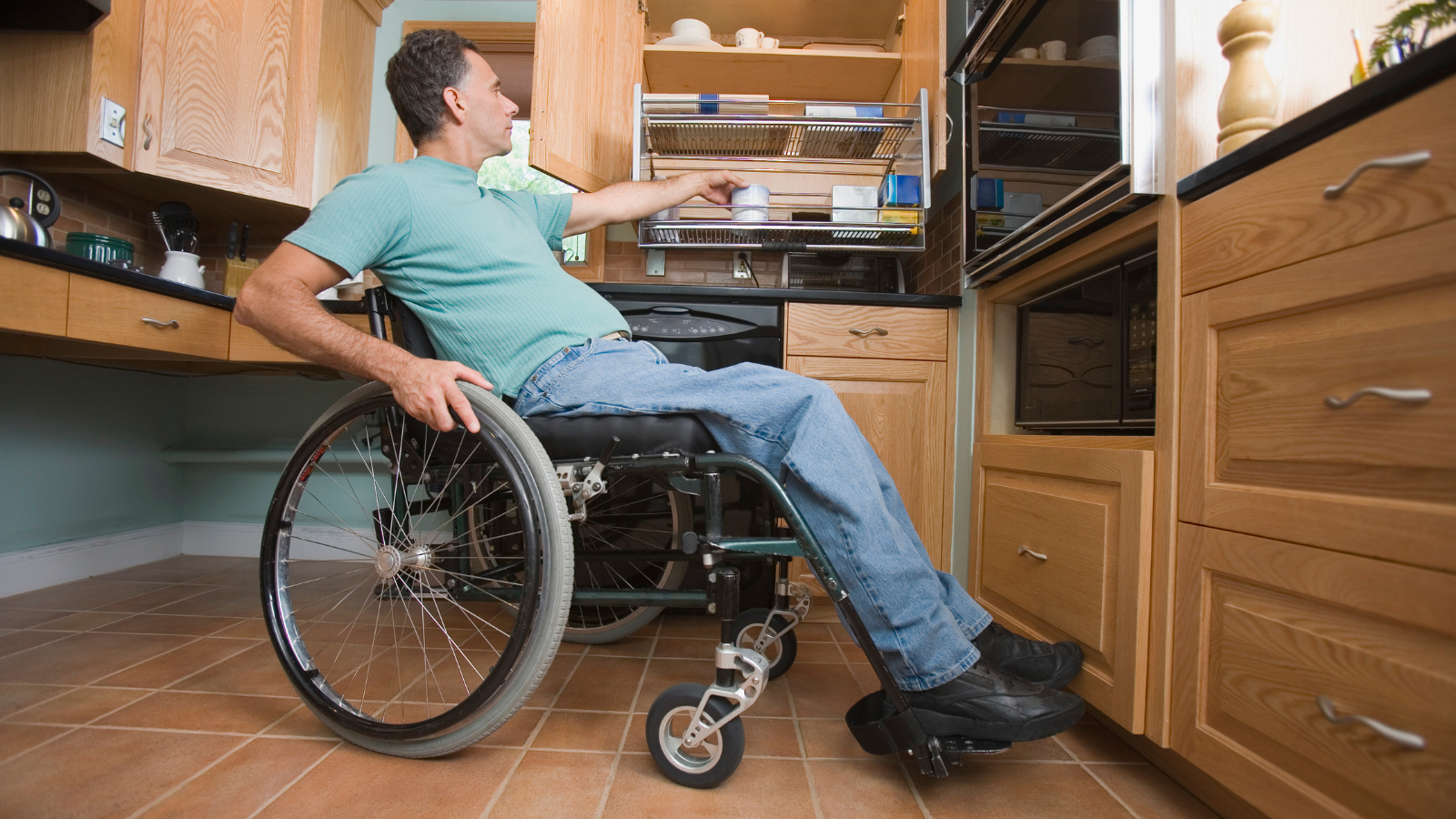 A man in a wheelchair is reaching into a cabinet in a kitchen.