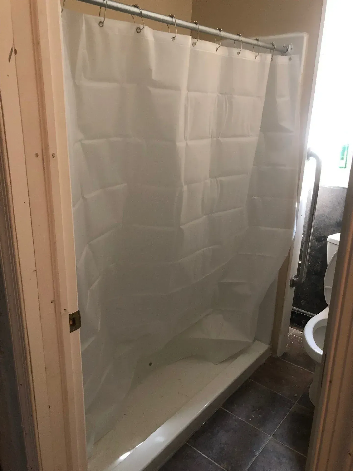 A shower stall with a white shower curtain in a bathroom next to a toilet.