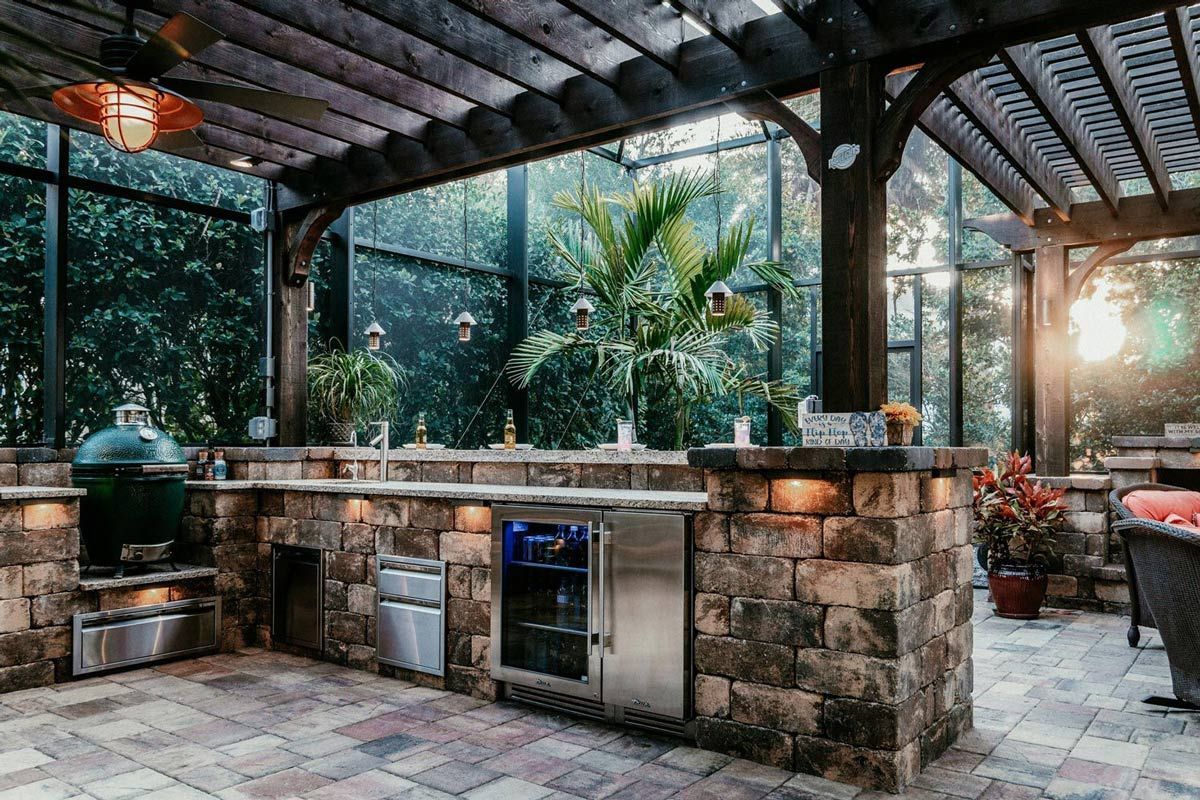 Outdoor stone patio kitchen with grill, sink, and bar under a covered roof, surrounded by greenery and sunlight