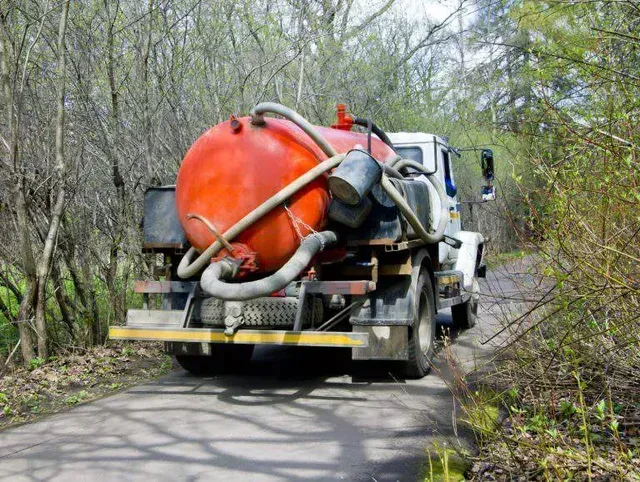 A white vacuum truck with a large, bright orange cylindrical tank driving along a narrow, tree-lined road.