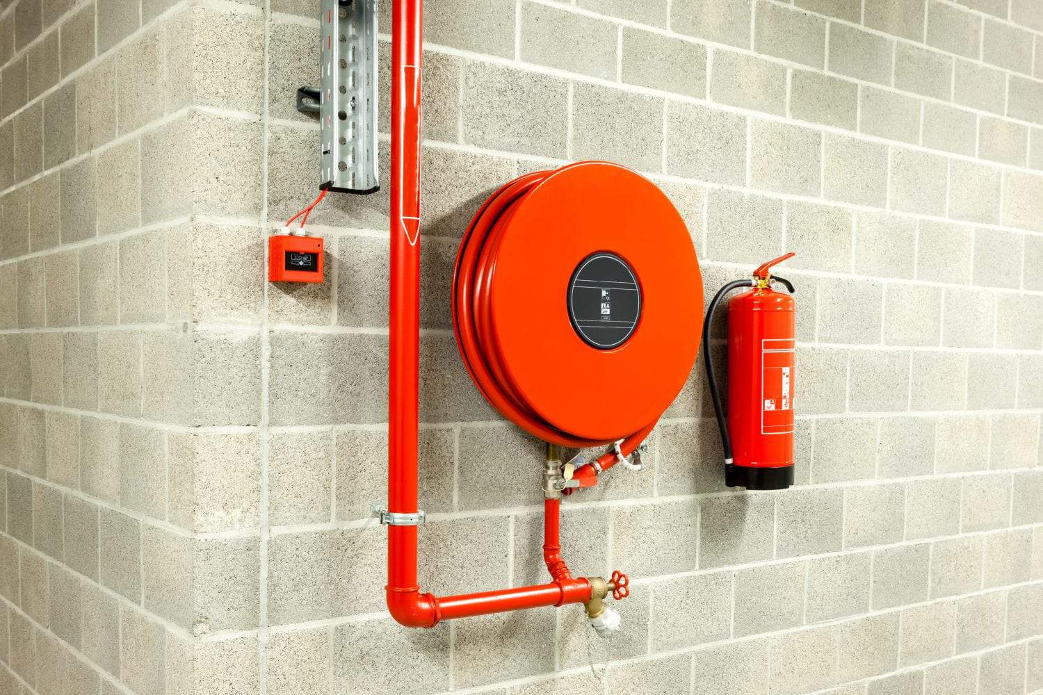 An orange fire hose reel and a red fire extinguisher mounted on a cinder block wall next to a red pipe.