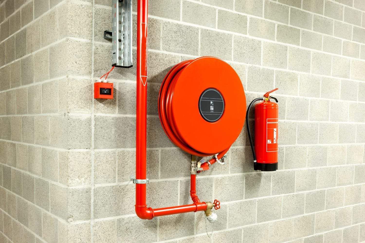 An orange fire hose reel and a red fire extinguisher mounted on a grey cinder block wall next to a vertical red pipe.