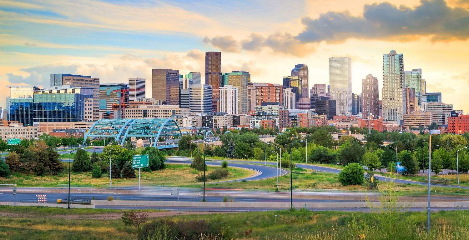 Denver skyline at sunset, featuring modern buildings, a distinctive blue bridge, and green space under a golden sky.