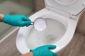 Hands in teal rubber gloves pouring cleaning powder from a measuring cup into a white toilet bowl.