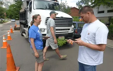 Three people stand on a street near a parked dump truck and several orange traffic cones; one person looks at a phone.