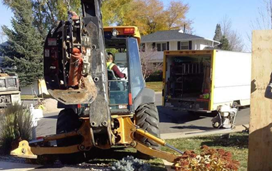 A backhoe parked on a residential street next to a utility truck with its rear doors open.