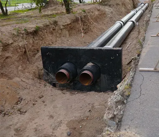 Two insulated, parallel metal pipes extend through a black support plate into an open trench along a roadside.