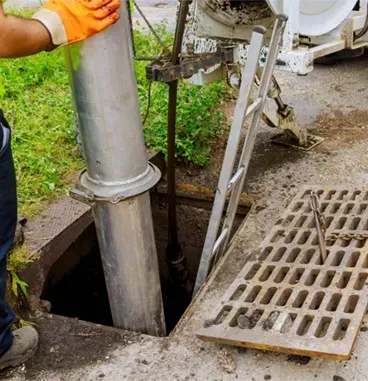 A worker in orange gloves uses a metal pipe to clear a storm drain grate located on a concrete sidewalk.