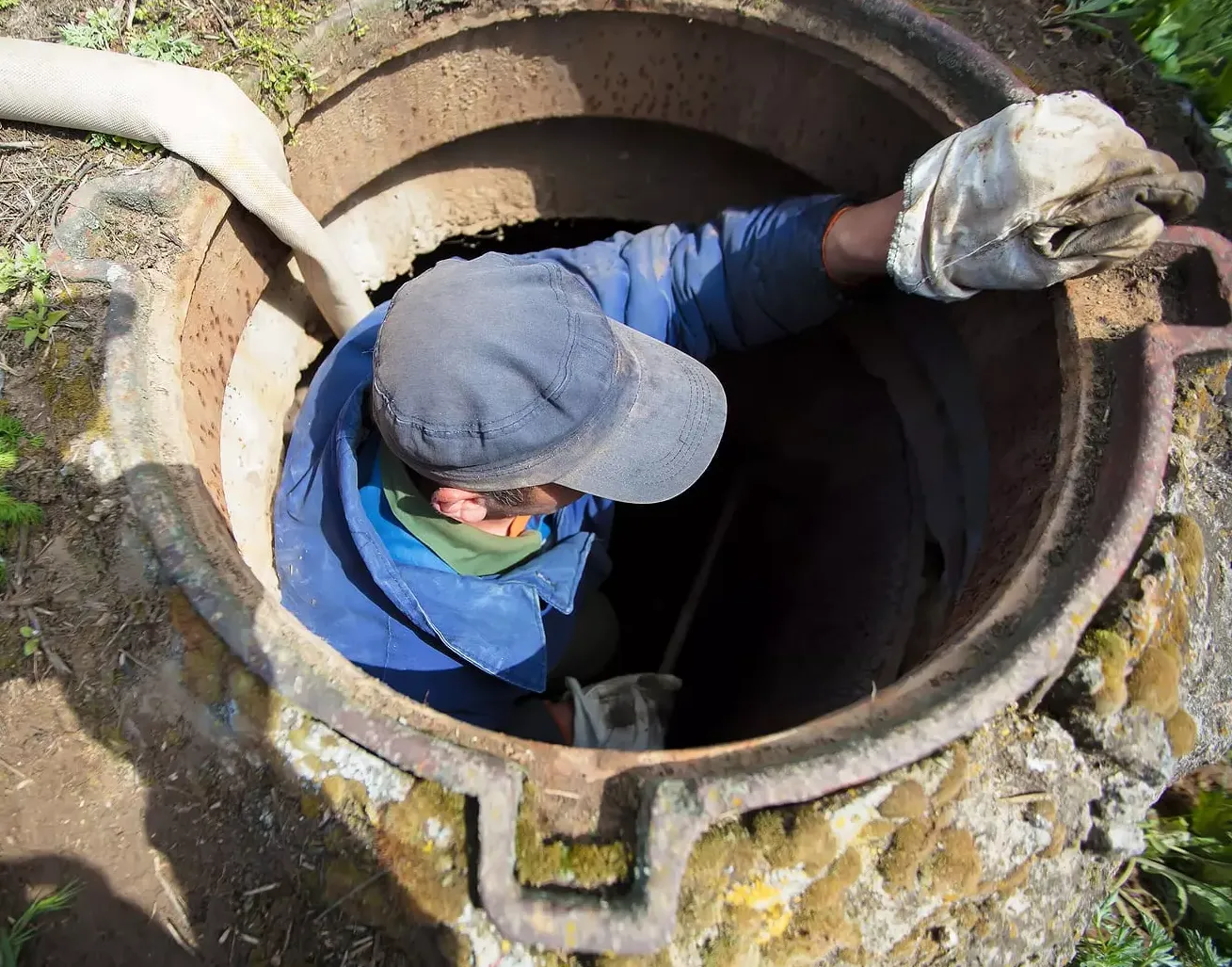 A person wearing a cap and gloves reaches into a deep, circular maintenance hole, with a hose nearby in an outdoor area.