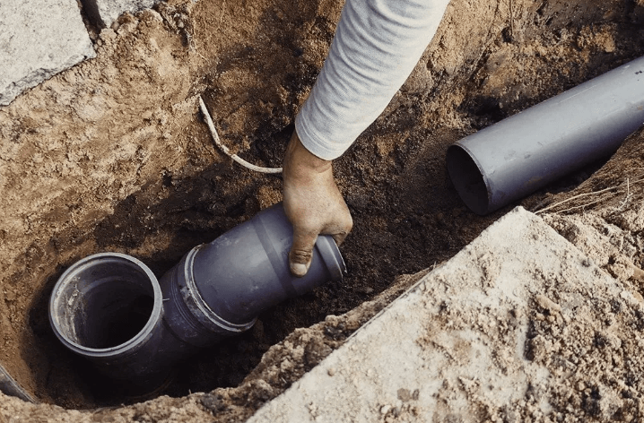 A person’s hand connecting gray plastic plumbing pipes in a narrow dirt trench.