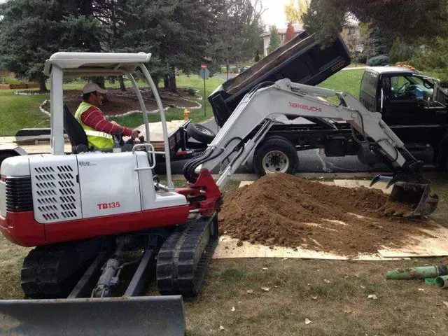 A worker operates a red and white mini excavator to pile dirt onto a truck on a residential lawn.