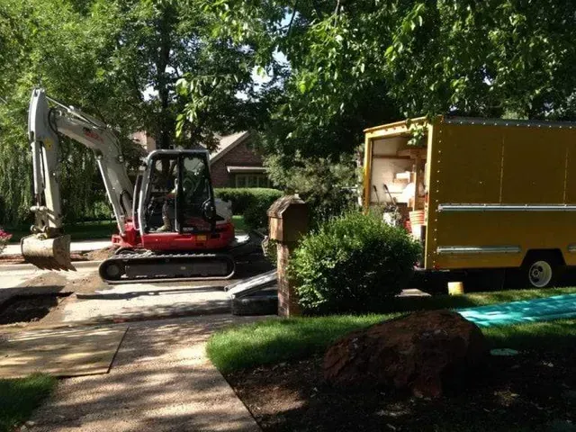 A construction excavator parked on a driveway next to a yellow utility truck in a wooded residential area.
